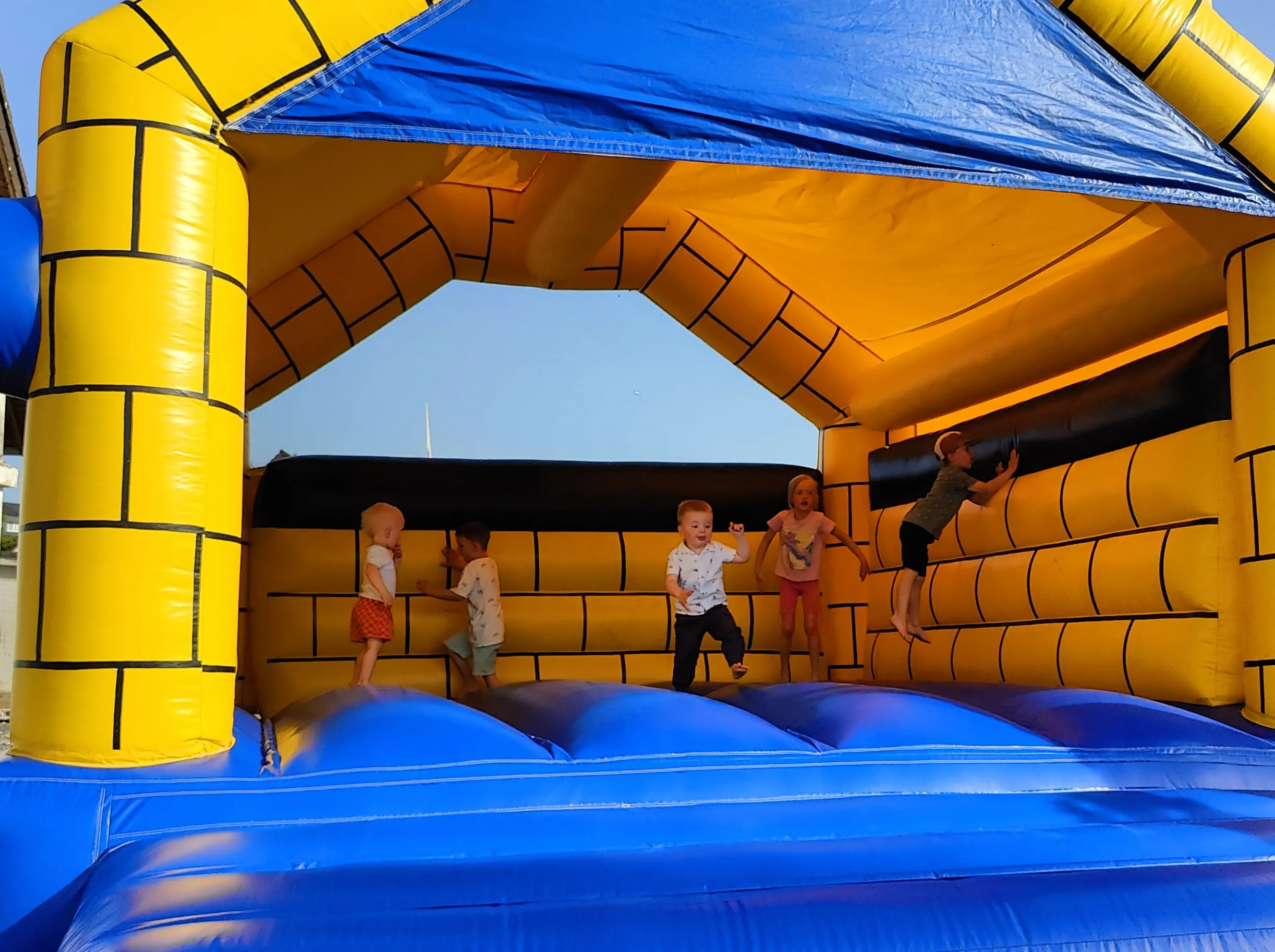 Mehrere Kinder spielen und klettern auf einer blauen und gelben Hüpfburg im Freien.