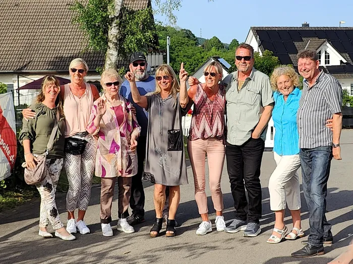 Eine Gruppe von acht lächelnden Menschen steht im Freien vor Häusern und Bäumen. Einige zeigen den Daumen nach oben.