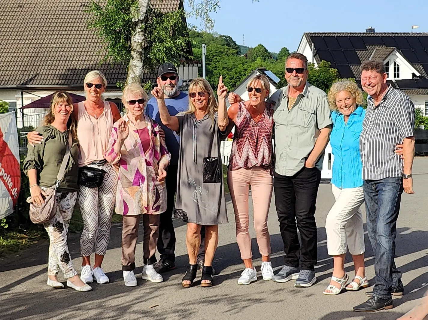 Eine Gruppe von acht lächelnden Menschen steht im Freien vor Häusern und Bäumen. Einige zeigen den Daumen nach oben.