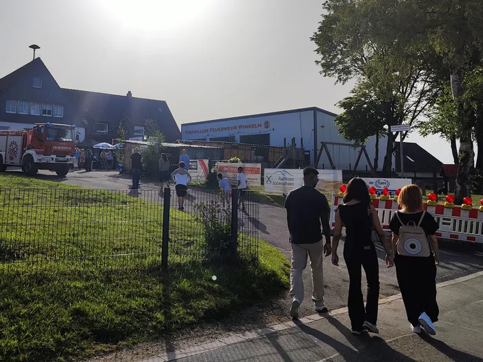 Drei Personen gehen auf eine Veranstaltung zu, vor der ein rotes Feuerwehrauto steht. Im Hintergrund ist das Gebäude der Freiwilligen Feuerwehr Winkeln zu sehen.
