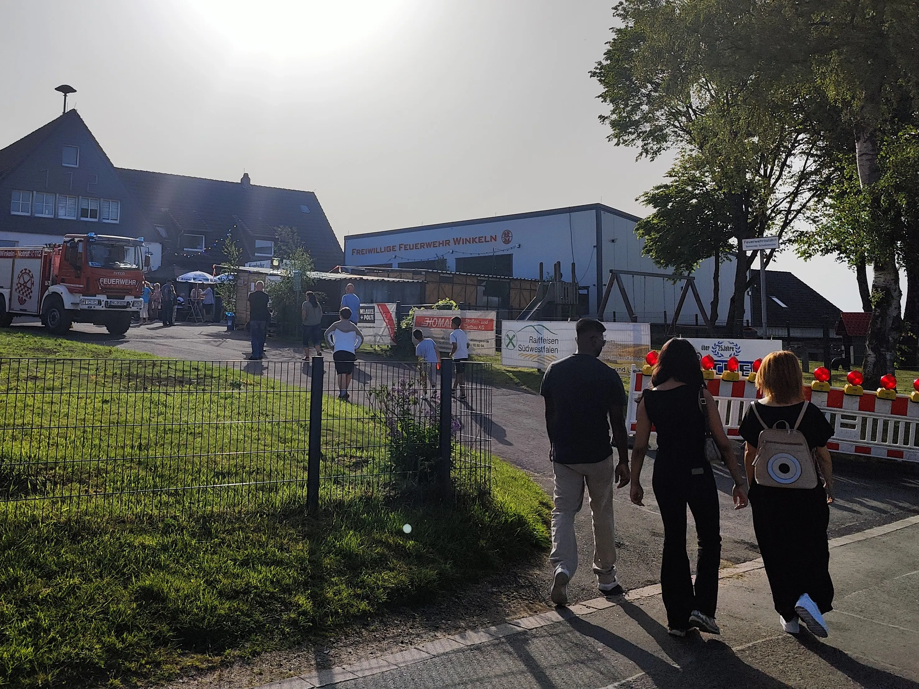 Drei Personen gehen auf eine Veranstaltung zu, vor der ein rotes Feuerwehrauto steht. Im Hintergrund ist das Gebäude der Freiwilligen Feuerwehr Winkeln zu sehen.