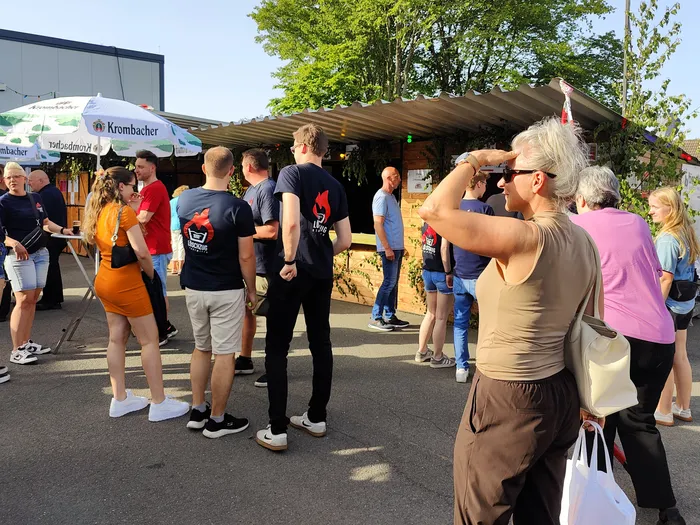 Menschen stehen vor einem Getränkestand mit dem Logo von „Krombacher“ auf einem Dorffest im Freien.