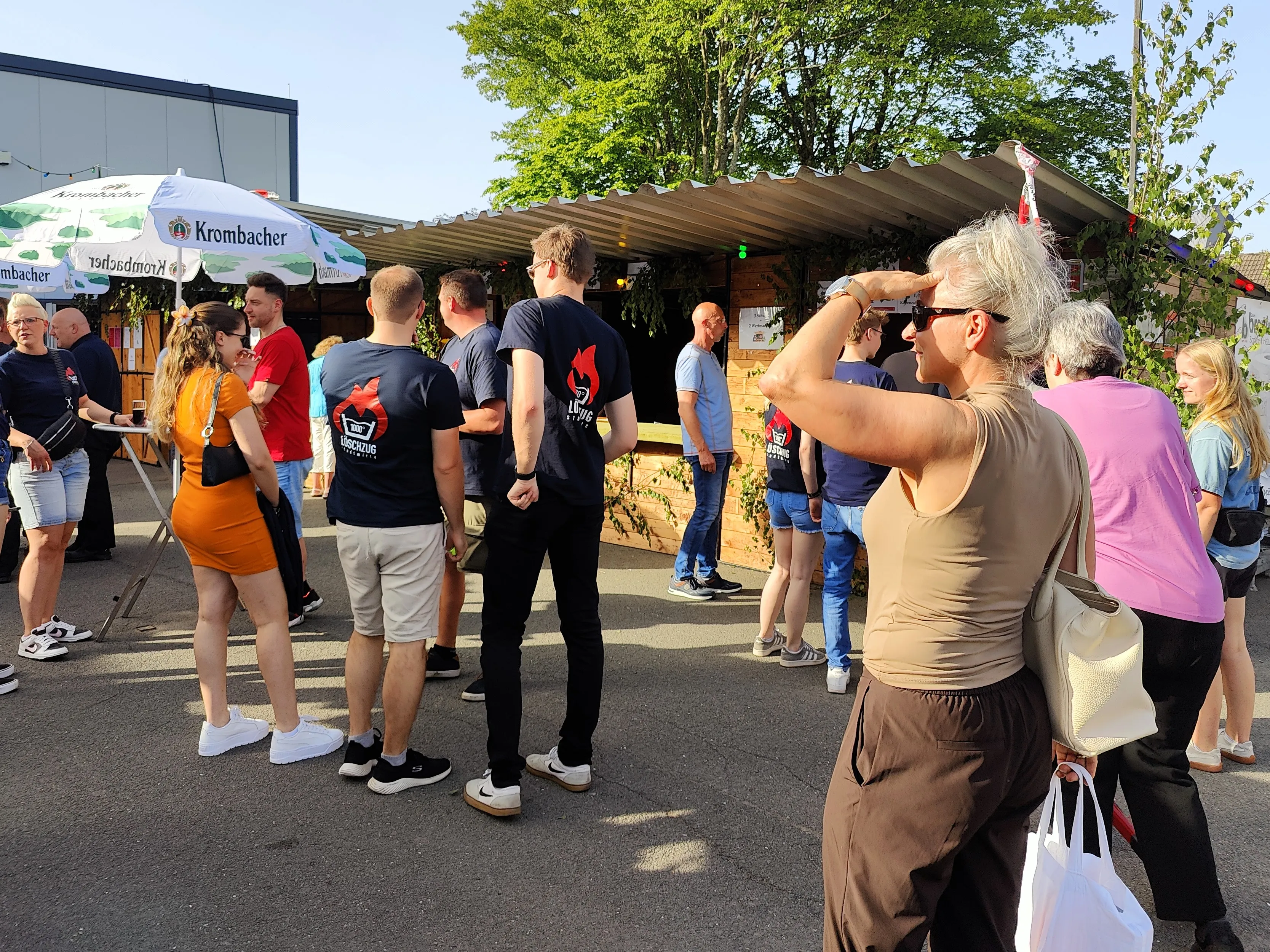 Menschen stehen vor einem Getränkestand mit dem Logo von „Krombacher“ auf einem Dorffest im Freien.