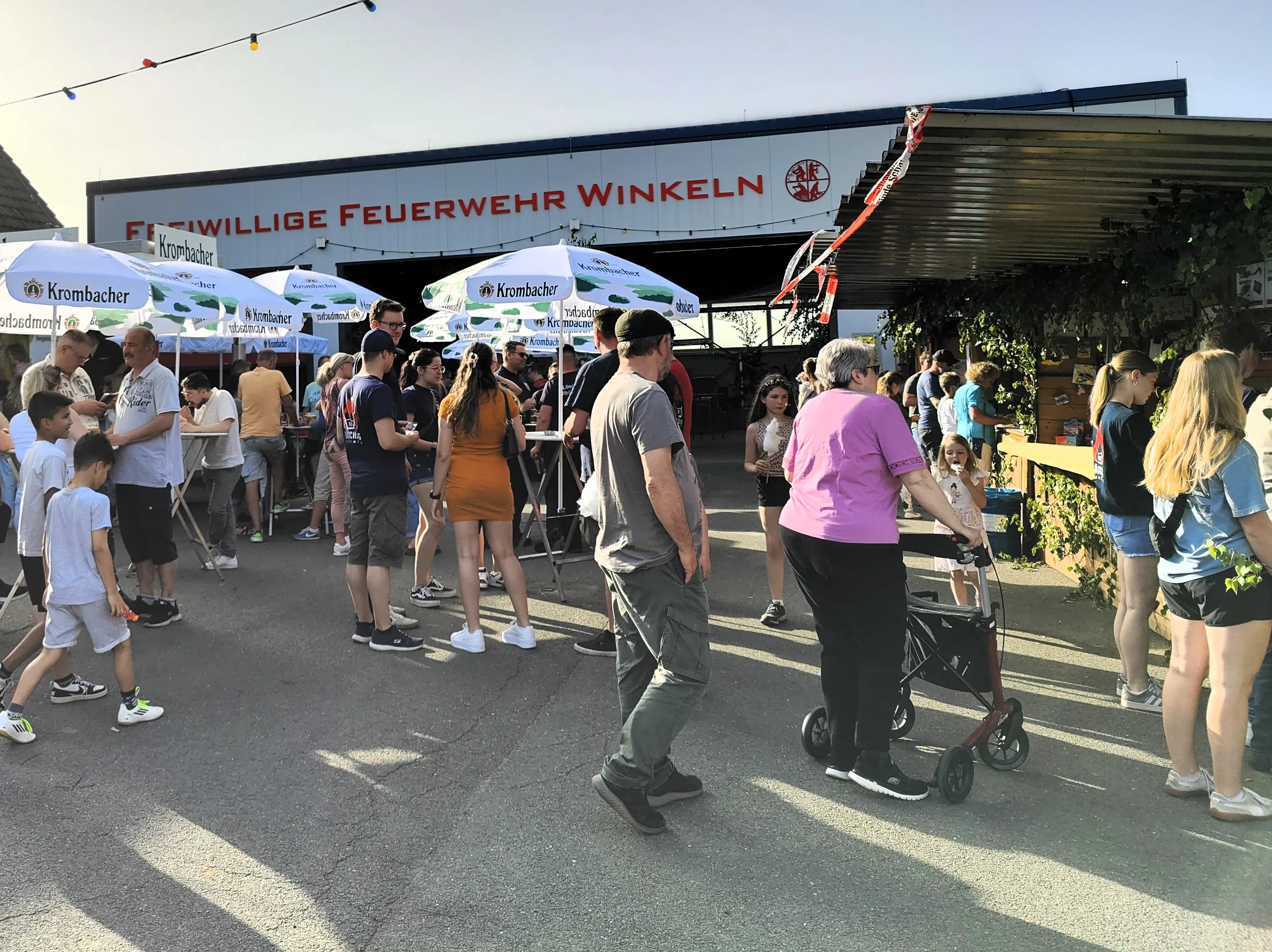 Menschengruppen stehen und gehen auf einem belebten Platz vor Zelten mit Werbeaufdrucken der Brauerei Krombacher. Im Hintergrund ein Schild mit der Aufschrift „Freiwillige Feuerwehr Winkeln“.
