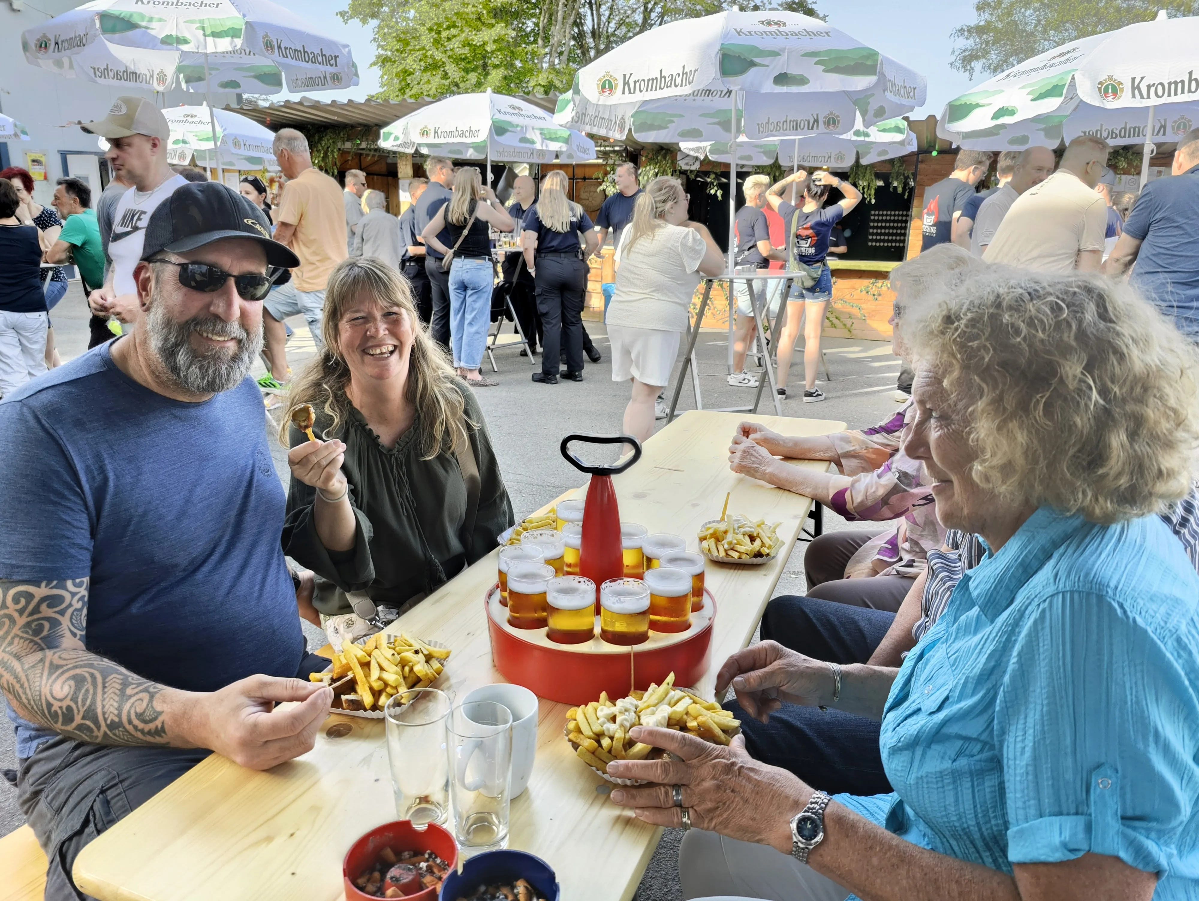 Drei Personen sitzen an einem Tisch im Freien und essen Pommes Frites. Im Hintergrund sind weitere Menschen und Schirme zu sehen.