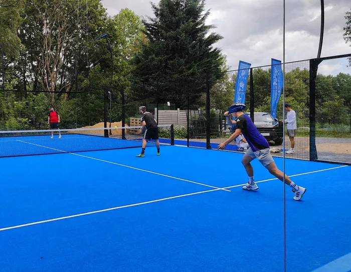 Drei Personen spielen auf einem hellblauen Pickleball-Platz, während eine vierte Person zuschaut. Der Spieler im Vordergrund schwingt einen Schläger, bereit, den Ball zu schlagen.