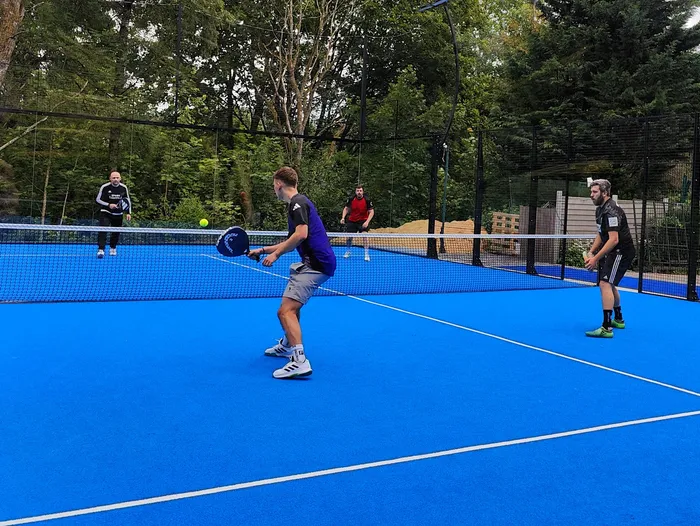 Ein Mann schwingt einen Schläger während eines Pickleball-Spiels auf einem blauen Platz. Im Hintergrund stehen weitere Spieler und ein schwarzes Drahtgeflecht.