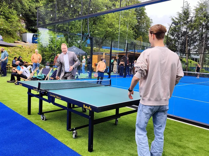 Ein Mann mit zurückgebundenen Haaren steht auf einem blauen Padel-Tennisplatz und blickt auf das Netz. Im Hintergrund stehen weitere Personen und ein Tischtennis-Tisch mit der Aufschrift 