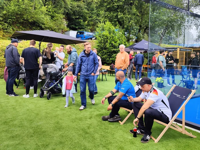 Menschen stehen und sitzen auf einer Rasenfläche vor einer Padel-Tennisanlage. Im Vordergrund sitzen zwei Männer in Sportkleidung auf Liegestühlen, während im Hintergrund ein Mann in Jeans und Jacke geht.