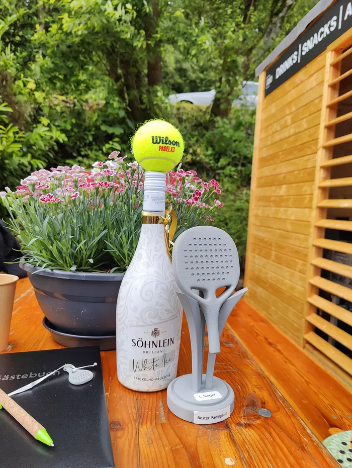 Eine Flasche Söhnlein White Ice steht neben einem Padel-Turnierpokal und einem Padelball auf einem Holztisch. Im Hintergrund ist ein Garten mit blühenden Pflanzen und ein Schild mit der Aufschrift 