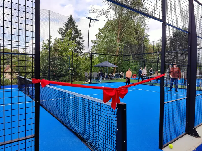 Ein Padel-Tennisplatz mit blauen Spielfeldern wird durch ein rotes Band eröffnet, umgeben von Zuschauern und Bäumen.