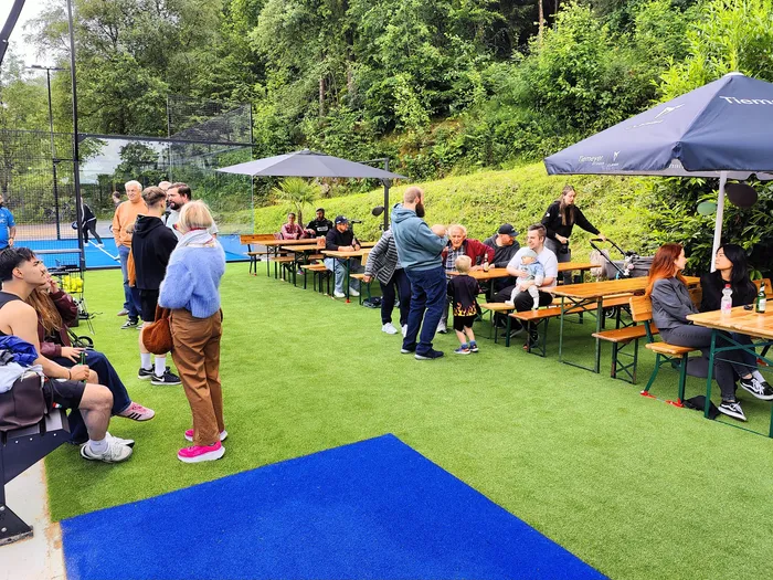 Menschen sitzen und stehen auf einer grünen Rasenfläche vor einer Padel-Tennisanlage. Einige Gäste sitzen an Biertischgarnituren unter Sonnenschirmen.