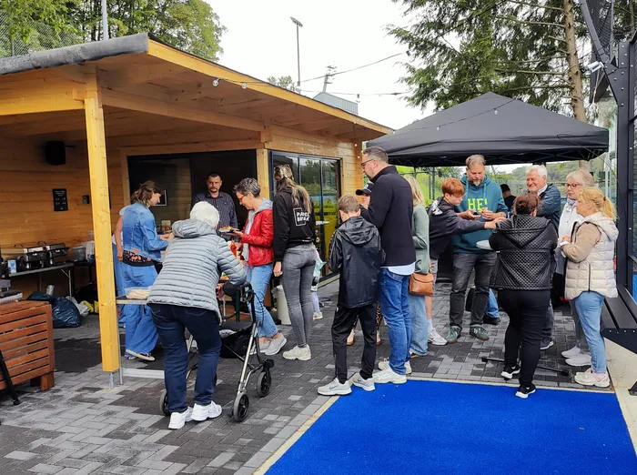 Eine Gruppe von Menschen steht vor einem Holzgebäude mit einem kleinen Vordach, vermutlich während der Eröffnung der 