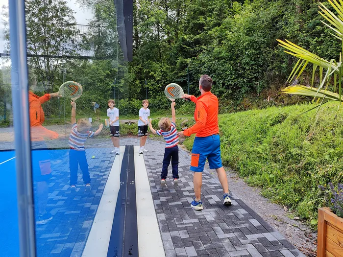 Ein Mann und drei Kinder spielen Padel auf einem Platz mit blauen und grauen Flächen. Im Hintergrund ist eine grüne Hecke und eine Palme zu sehen.