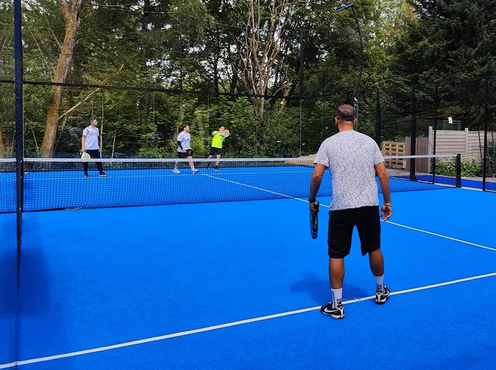 Ein Spieler steht mit dem Rücken zum Betrachter auf einem blauen Pickleball-Platz und hält einen Schläger. Im Hintergrund sind drei weitere Spieler zu sehen, die bereit für das Spiel stehen.