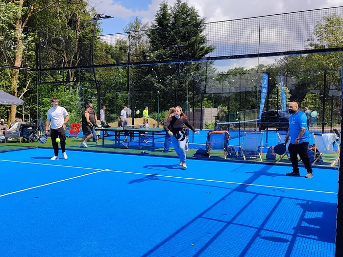 Mehrere Personen spielen auf einem blauen Pickleball-Platz. Eine Frau in Jogginghose bereitet einen Schlag vor, während ein Mann mit einem Schläger bereit steht. Im Hintergrund sind weitere Spieler und Zuschauer zu sehen.