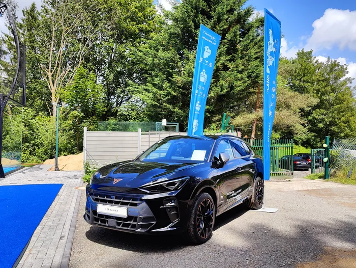 Dunkler Cupra Terramar steht auf einer blauen Fläche vor grünem Hintergrund mit Bäumen und Bannern. Das Auto ist das Hauptmotiv und wird vermutlich von den Gewinnern eines Gewinnspiels genutzt.