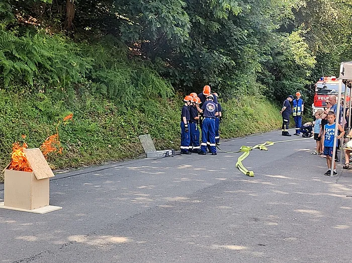 Feuerwehrleute in blauer Arbeitskleidung und orangefarbenen Westen kontrollieren ein kleines Feuer in einem Holzkasten auf einer Landstraße. Im Hintergrund steht ein Feuerwehrauto und einige Zuschauer beobachten die Übung.