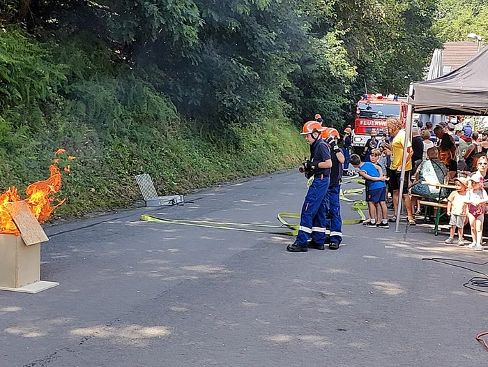 Zwei Feuerwehrleute mit Helmen und Schutzkleidung stehen vor einem brennenden Container und halten einen Schlauch bereit. Im Hintergrund stehen Zuschauer und ein rotes Feuerwehrauto.