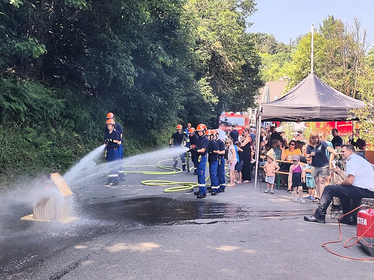Feuerwehrleute löschen mit Wasserstrahlen ein brennendes Objekt während eines Tages der offenen Tür. Im Hintergrund stehen Zuschauer unter einem Zelt und ein Feuerwehrauto ist zu sehen.
