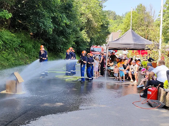 Feuerwehrleute demonstrieren vor einer Zuschauermenge die Brandbekämpfung mit einem Wasserstrahl auf ein kleines Holzhaus. Im Hintergrund steht ein Zelt und ein Feuerwehrauto.