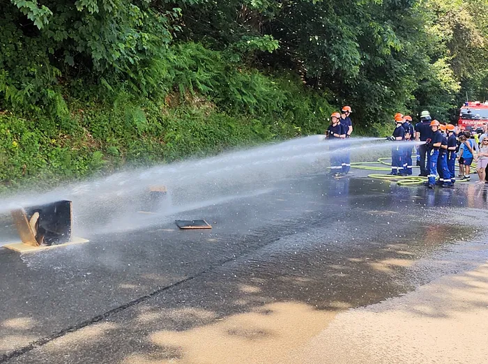 Feuerwehrleute löschen mit einem Wasserstrahl eine Brandübung auf einem asphaltierten Platz. Im Hintergrund stehen weitere Einsatzkräfte und zusehen, während Kinder und Erwachsene zuschauen.
