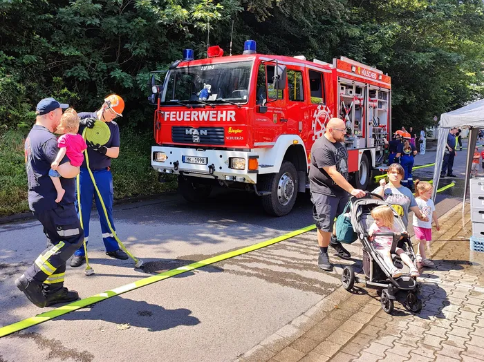 Feuerwehrleute in Uniform stehen vor einem roten Feuerwehrauto der Marke MAN, während im Hintergrund Besucher eines Tages der offenen Tür zu sehen sind. Ein Kind sitzt in einem Buggy, während ein anderer Mensch einen Kinderwagen schiebt.