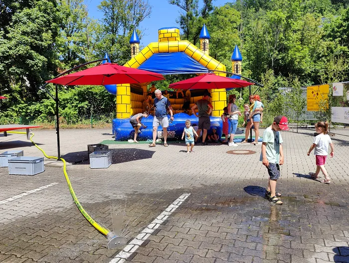 Children play in and around a blue and yellow bouncy castle with a castle theme during an open house event at the Schalksmühle fire department. Adults stand around the castle, and a water hose sprays water onto the ground nearby.