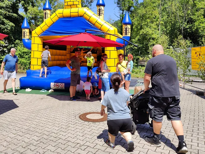 People gather around a large, colorful inflatable castle at an outdoor event. A person in a bear costume is visible inside the castle, and several adults and children are watching.