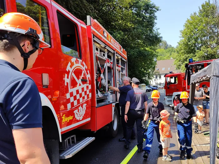 Rotes Feuerwehrauto mit aufwendiger Grafik an der Seite steht im Mittelpunkt eines Tages der offenen Tür. Feuerwehrleute und Besucher, darunter auch Kinder mit Helmen, bewegen sich um das Fahrzeug herum.