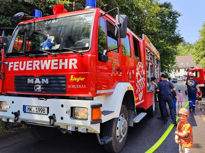 A bright red MAN fire engine with 'FEUERWEHR' prominently displayed on the side. A child in a colorful outfit stands near the truck, looking towards the camera, while firefighters stand nearby during an open house event.