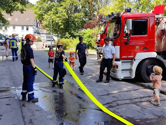 Feuerwehrleute demonstrieren Kindern die Handhabung eines gelben Schlauchs vor einem roten Feuerwehrauto mit der Aufschrift 