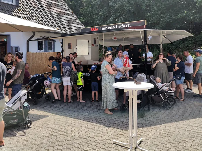 Eine Menschenmenge steht vor einem Imbisswagen mit der Aufschrift „linnepe liefert.“ am Tag der offenen Tür der Feuerwehr in Schalksmühle. Einige Besucher halten Kinder im Arm oder haben Kinderwagen dabei.
