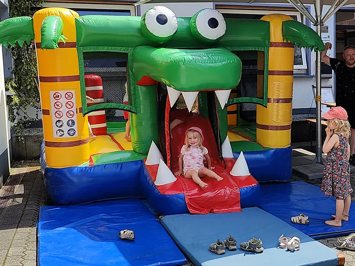 A colorful inflatable crocodile serves as a bouncy castle with a slide at a fire department's open day. A young child is sliding down, while another child stands ready to climb up.