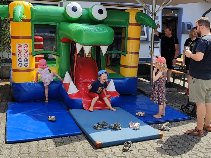 Kinder spielen vor einer aufblasbaren Krokodil-Hüpfburg auf dem Tag der offenen Tür der Feuerwehr Schalksmühle. Schuhe liegen verstreut vor dem Eingang. Im Hintergrund stehen Erwachsene und ein Tisch mit Sonnenschirm.