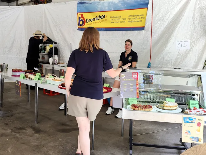 A woman with blonde hair wearing a blue polo shirt and yellow pants is standing at a food stand with cakes and pastries. In the background, a firefighter stands near a tent with the 'Village on Fire' logo and contact information for 'bremicker'.