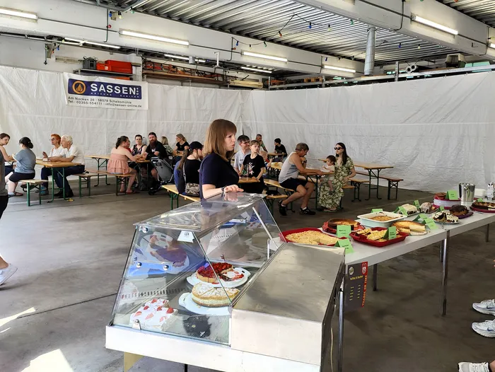 A woman stands behind a table displaying cakes and pastries at an event with people seated at long tables in the background. A sign for 