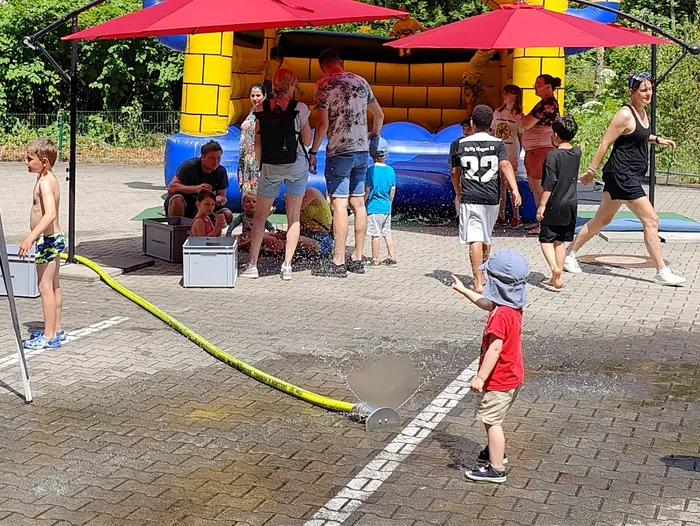 Kinder spielen mit einem Wasserschlauch auf einem gepflasterten Platz vor einer Hüpfburg, während im Hintergrund Erwachsene stehen und zusehen. Im Hintergrund sind Sonnenschirme und Bäume zu sehen.