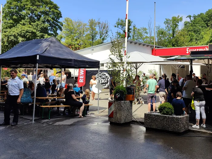 People are gathered at a village fair with food and drink stalls, including a Feuerwehr (fire department) stand, on a sunny day. Several people are seated at picnic tables under a dark blue canopy, while others stand in line at a food stall.