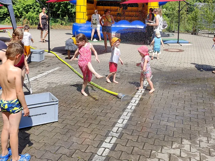 Kinder spielen auf dem Tag der offenen Tür der Feuerwehr Schalksmühle mit Wasser. Ein Junge hält einen Wasserschlauch, während andere Kinder damit bespritzt werden. Im Hintergrund ist eine Hüpfburg und weitere Besucher zu sehen.