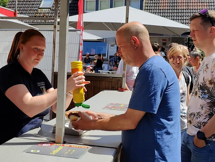 A woman in a black shirt is applying mustard to a sausage roll for a man with a shaved head and a blue t-shirt at an outdoor food stall. In the background, people are gathered at a fire department open day.