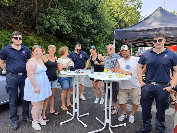 Eine Gruppe von Menschen steht um einen Tisch mit Getränken und Snacks herum. Im Hintergrund ist ein rotes Feuerwehrfahrzeug und ein Zelt zu sehen.