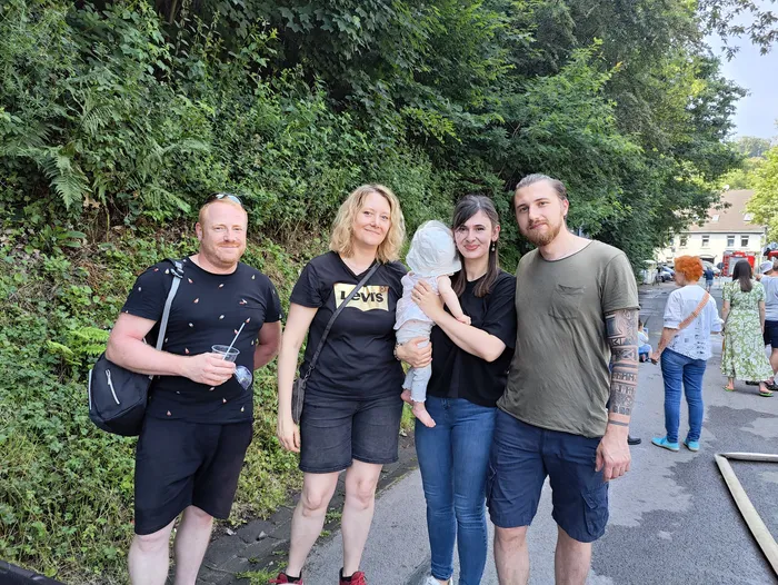 A group of four adults and a baby stand closely together, smiling at the camera. Behind them, people are milling about at an outdoor event with green foliage and buildings visible in the background.