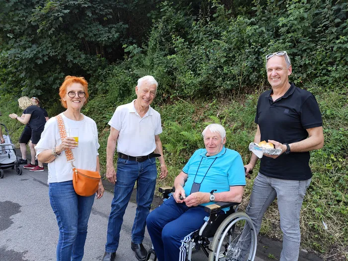 Four people pose for a photo at an event. A woman with red hair and a man are standing, while another man sits in a wheelchair. A fourth man holds a plate with food. In the background, people are gathered near a road, and lush greenery is visible.