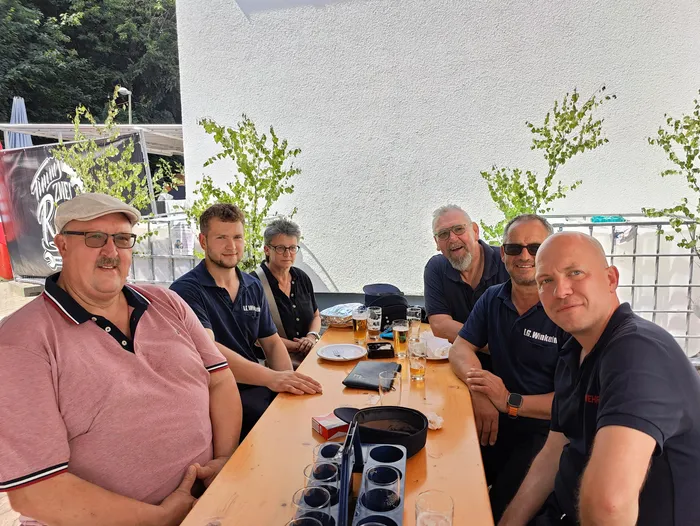 Sechs Männer sitzen an einem Holztisch im Freien, vor ihnen stehen Biergläser und Snacks. Sie tragen Freizeitkleidung, einige haben Sonnenbrillen auf und lächeln in die Kamera. Im Hintergrund ist ein Gebäude und ein Baum zu sehen.