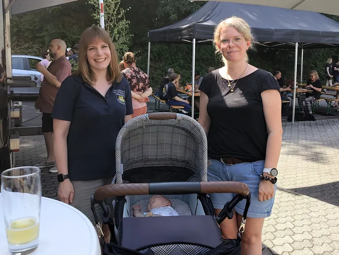 Zwei Frauen stehen lächelnd neben einem Kinderwagen, in dem ein Baby liegt. Im Hintergrund sind Menschen und ein Feuerwehrauto zu sehen, während ein Pavillon für Schatten sorgt.