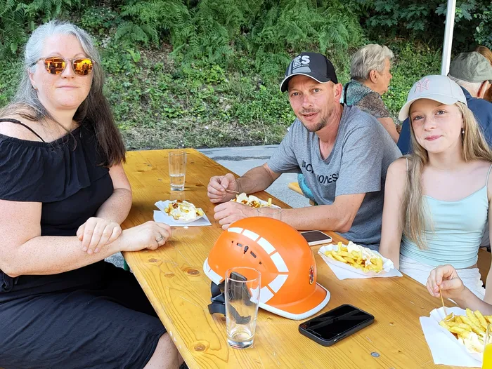 A group of people sits around a wooden picnic table enjoying food. A bright orange firefighter's helmet is prominently placed on the table alongside smartphones and drinks.