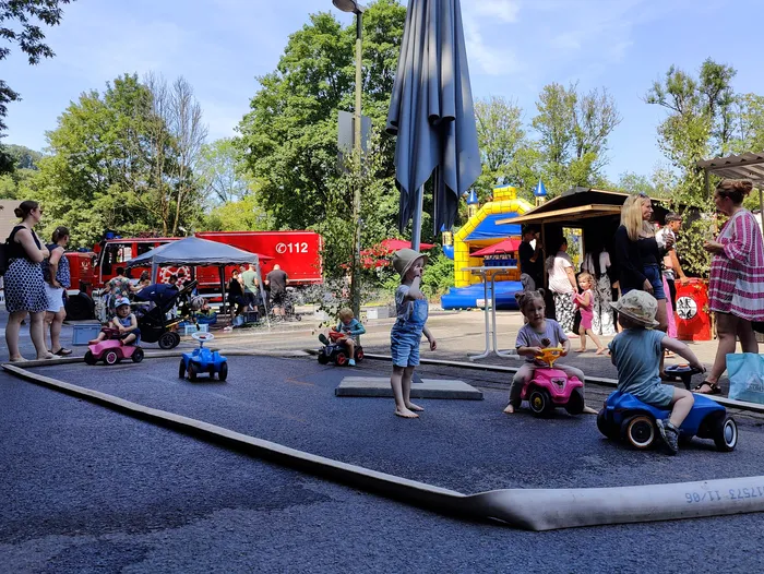 Kinder fahren mit kleinen Elektroautos auf einem Platz vor einer Feuerwache, während im Hintergrund eine Hüpfburg steht und Menschen zusehen.