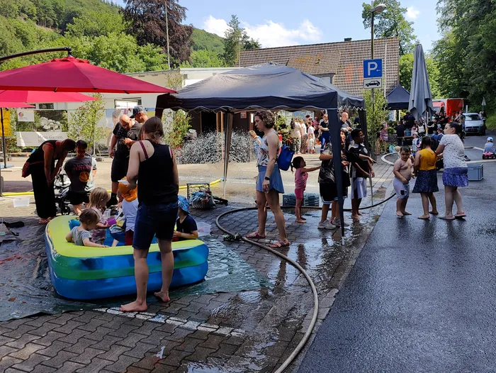 Kinder planschen und spielen unter einem Wasserstrahl, der von Feuerwehrleuten während eines Tages der offenen Tür in Schalksmühle erzeugt wird. Im Hintergrund sind ein Zelt, Menschen und ein Parkplatzschild zu sehen.
