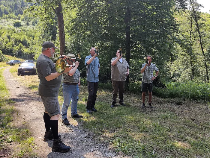 Fünf Männer spielen im Wald Blasinstrumente, darunter ein Tenorhorn. Im Hintergrund sind Bäume und geparkte Autos zu sehen.